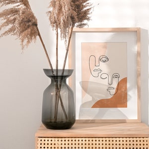 Puede incluir: Un jarr&oacute;n de vidrio gris oscuro con hierba de la pampa seca sobre una mesa de madera con una impresi&oacute;n enmarcada de caras de arte abstracto de l&iacute;neas en tonos marrones y beige.