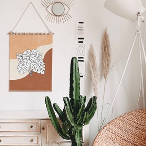 May include: A white wall with a brown and orange framed print of a white leaf design. The print is hanging on a wall with a wooden dowel rod. There is a large green cactus plant in front of the print. The wall also has a gold mirror with a sunburst design and a white floor lamp with a white shade.