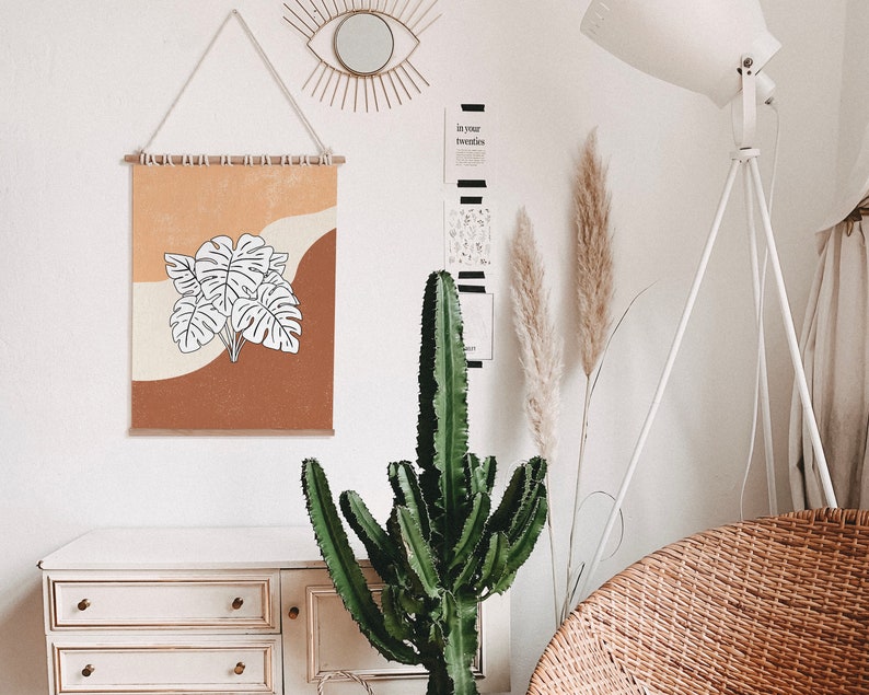 May include: A white wall with a brown and orange framed print of a white leaf design. The print is hanging on a wall with a wooden dowel rod. There is a large green cactus plant in front of the print. The wall also has a gold mirror with a sunburst design and a white floor lamp with a white shade.