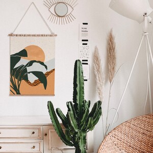 May include: A white dresser with three drawers, a green cactus plant, a white lamp with a white shade, and a wall hanging with a sunset and palm trees design. The wall hanging is made of fabric and has a wooden frame.
