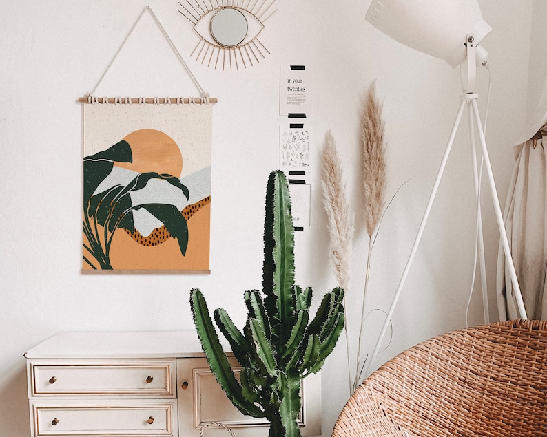 May include: A white dresser with three drawers, a green cactus plant, a white lamp with a white shade, and a wall hanging with a sunset and palm trees design. The wall hanging is made of fabric and has a wooden frame.