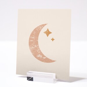 May include: A beige card with a pink crescent moon and two gold stars. The moon has a textured look.