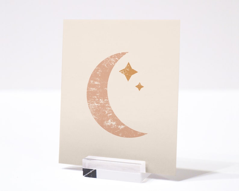 May include: A beige card with a pink crescent moon and two gold stars. The moon has a textured look.