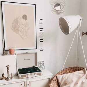 May include: A white record player with a gold trim sits on a white dresser with a candle and candle holder. A framed print with a line drawing of flowers on a beige background hangs on the wall above the dresser. A white lamp with a tripod stand is in the background.