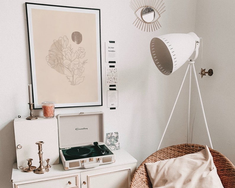 May include: A white record player with a gold trim sits on a white dresser with a candle and candle holder. A framed print with a line drawing of flowers on a beige background hangs on the wall above the dresser. A white lamp with a tripod stand is in the background.