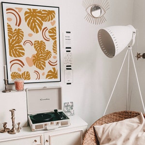 May include: A white record player with a black turntable and gold accents sits on a white dresser with a framed print of a brown and orange abstract design with monstera leaves. A white lamp with a tripod stand is in the background.
