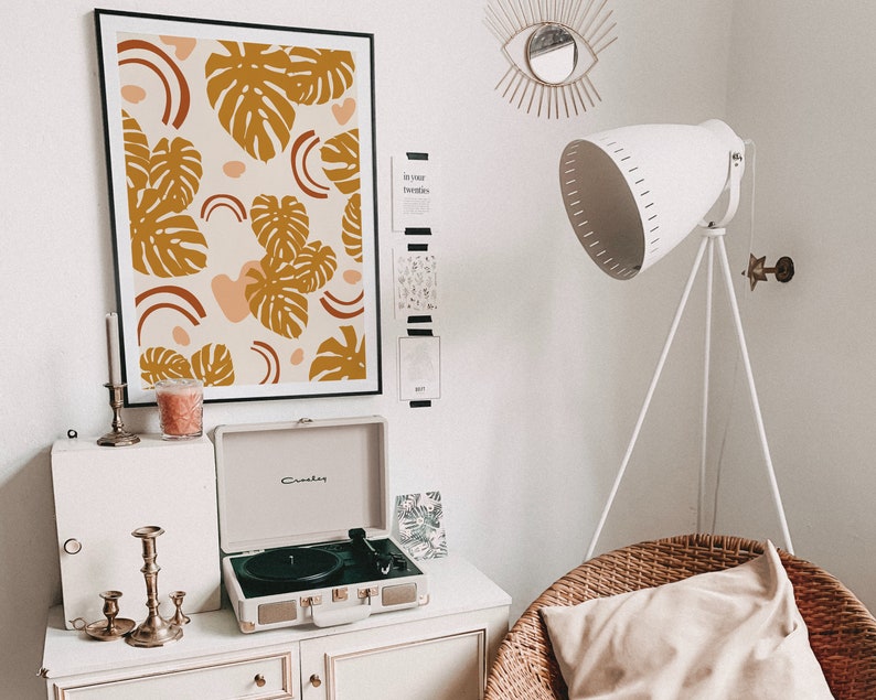 May include: A white record player with a black turntable and gold accents sits on a white dresser with a framed print of a brown and orange abstract design with monstera leaves. A white lamp with a tripod stand is in the background.