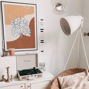 May include: A white record player with a brown and orange framed print of a tropical leaf design above it. The print is on a white wall with a gold eye-shaped mirror and a white lamp with a tripod stand.
