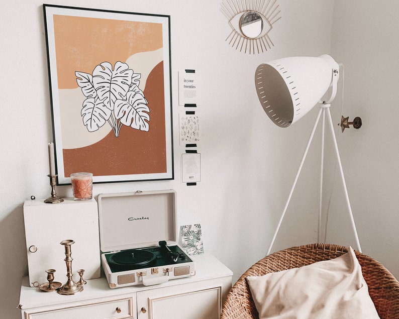 May include: A white record player with a brown and orange framed print of a tropical leaf design above it. The print is on a white wall with a gold eye-shaped mirror and a white lamp with a tripod stand.