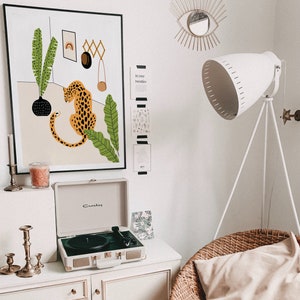 May include: A white record player with a black turntable and gold accents sits on a white shelf with a candle and candle holders. A framed print of a leopard with green leaves hangs on the wall above the record player. The print has the text "In your twenties" on the wall next to it.