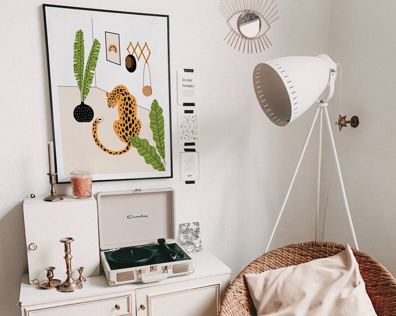 May include: A white record player with a black turntable and gold accents sits on a white shelf with a candle and candle holders. A framed print of a leopard with green leaves hangs on the wall above the record player. The print has the text "In your twenties" on the wall next to it.