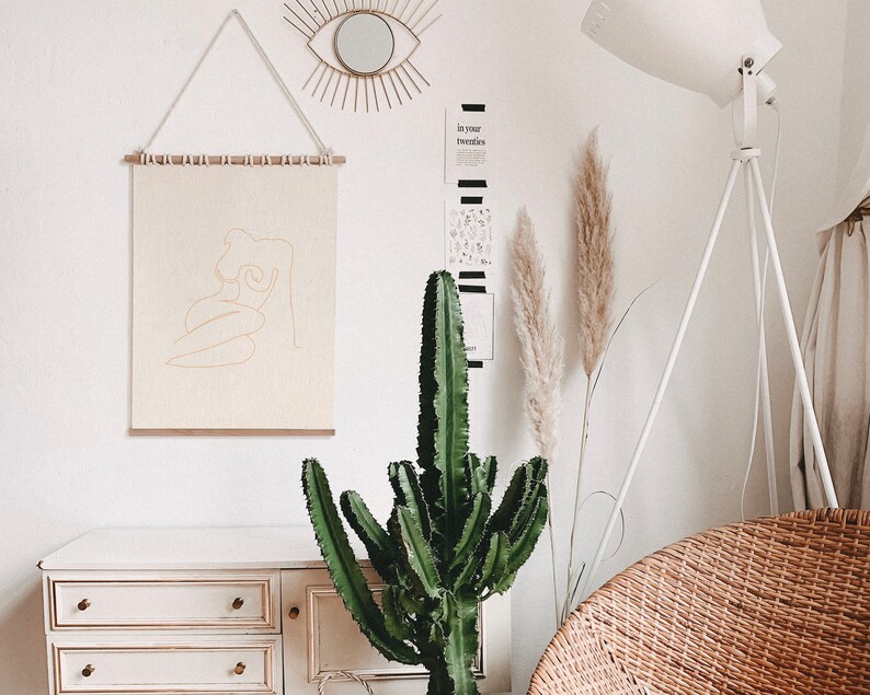 May include: A white wall with a beige canvas print of a line drawing of a woman. The print is hanging on a wooden dowel rod with white string. A large green cactus plant is in front of a white dresser with three drawers. A white floor lamp with a white shade is in the background. The text "in your twenties" is on a black and white card on the wall.