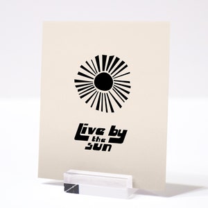 Puede incluir: Una impresi&oacute;n en blanco y negro con un dise&ntilde;o de sol estilizado y el texto "Live by the Sun".