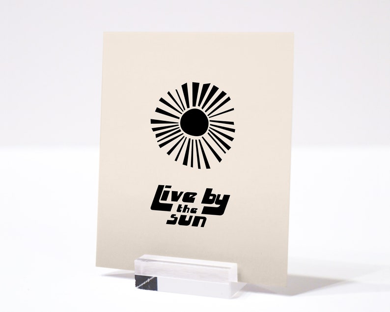 Puede incluir: Una impresi&oacute;n en blanco y negro con un dise&ntilde;o de sol estilizado y el texto "Live by the Sun".