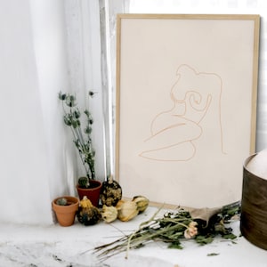 May include: A framed line drawing of a nude figure in a seated pose. The drawing is in a light brown color and is on a beige background. The frame is a light wood color.