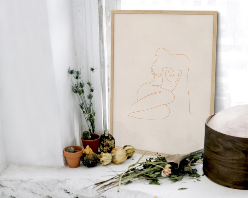 May include: A framed line drawing of a nude figure in a seated pose. The drawing is in a light brown color and is on a beige background. The frame is a light wood color.
