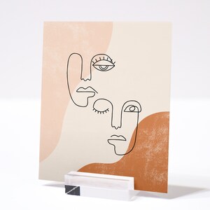 Puede incluir: Impresi&oacute;n de arte abstracto de l&iacute;nea de tres caras con un fondo melocot&oacute;n y marr&oacute;n. Las caras est&aacute;n dibujadas en l&iacute;neas negras y tienen rasgos simples. La impresi&oacute;n es perfecta para agregar un toque moderno a cualquier habitaci&oacute;n.