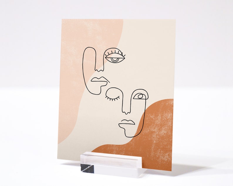 Puede incluir: Impresi&oacute;n de arte abstracto de l&iacute;nea de tres caras con un fondo melocot&oacute;n y marr&oacute;n. Las caras est&aacute;n dibujadas en l&iacute;neas negras y tienen rasgos simples. La impresi&oacute;n es perfecta para agregar un toque moderno a cualquier habitaci&oacute;n.