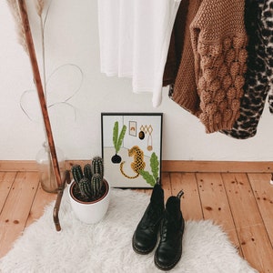 May include: A framed print of a jungle scene with a leopard, plants, and a rainbow. The print is hanging on a white wall with a copper pipe rack in the background. There are two pairs of black boots on a white fluffy rug in the foreground.