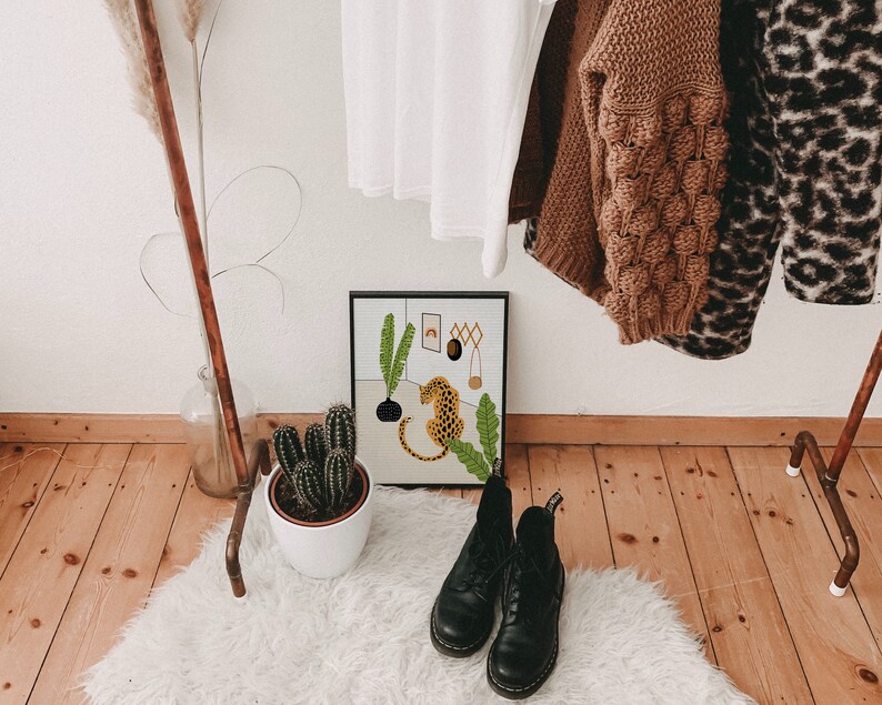 May include: A framed print of a jungle scene with a leopard, plants, and a rainbow. The print is hanging on a white wall with a copper pipe rack in the background. There are two pairs of black boots on a white fluffy rug in the foreground.