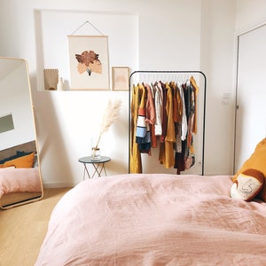May include: A bedroom with a pink linen duvet cover, a black clothing rack with a variety of clothes hanging on it, a full-length mirror, and a small side table with a vase of dried pampas grass.
