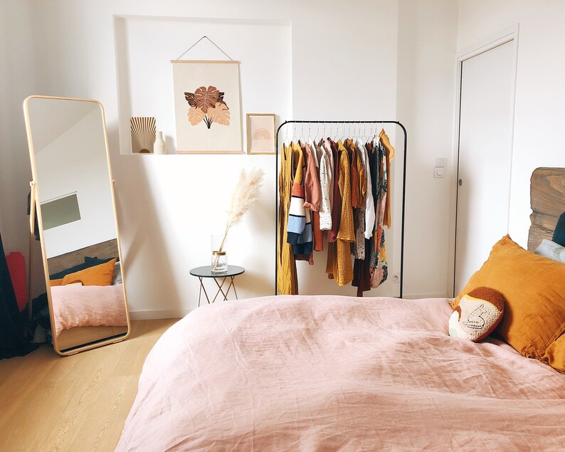 May include: A bedroom with a pink linen duvet cover, a black clothing rack with a variety of clothes hanging on it, a full-length mirror, and a small side table with a vase of dried pampas grass.