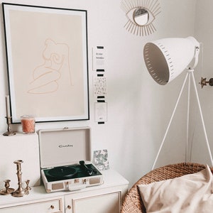 May include: A white record player with a gold trim sits on a white dresser with candles and a framed line drawing of a woman in a beige frame. A white lamp with a tripod stand is in the background.