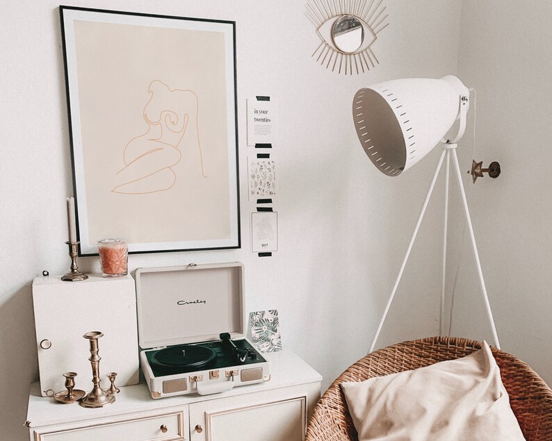 May include: A white record player with a gold trim sits on a white dresser with candles and a framed line drawing of a woman in a beige frame. A white lamp with a tripod stand is in the background.