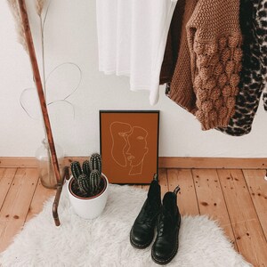 May include: A black framed print with a minimalist line drawing of two faces in an orange background. The print is hanging on a white wall with a wooden rack above it. The rack is holding a brown sweater, a leopard print sweater, and a white shirt.