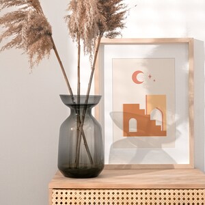 May include: A dark gray glass vase with dried pampas grass sits on a light wood table next to a framed print of a geometric building with a crescent moon and stars.