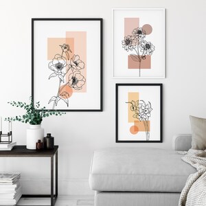 May include: Three black framed prints with white line drawings of flowers on a peach background. The prints are hung on a white wall.