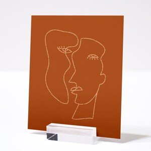 May include: A minimalist line art print of two faces on a burnt orange background. The faces are drawn in a single line and are overlapping.