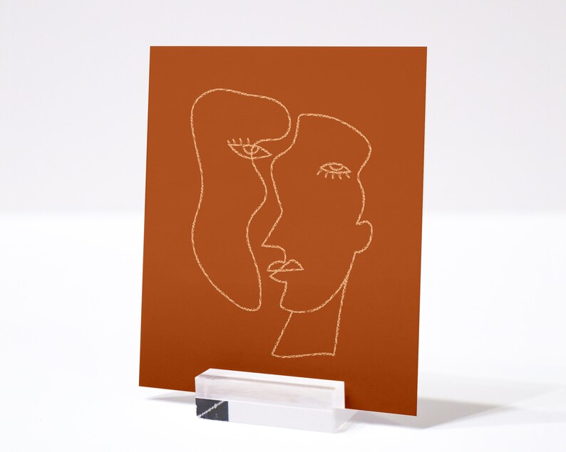May include: A minimalist line art print of two faces on a burnt orange background. The faces are drawn in a single line and are overlapping.