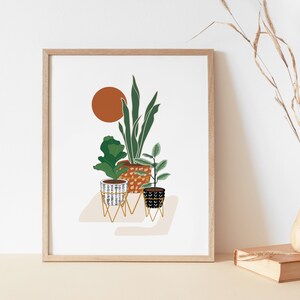 以下が含まれることがあります： 幾何学模様の鉢植えの植物が3つ描かれた、ミニマルなイラストの額装プリント。植物は、白い背景に茶色の太陽が描かれた左上の隅に、金色の金属製のスタンドの上に置かれている。