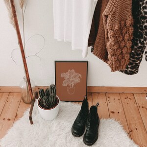 May include: A brown framed print with a line drawing of a palm tree in a pot on a terracotta background. The print is hanging on a white wall above a white fluffy rug with two black leather boots on it.