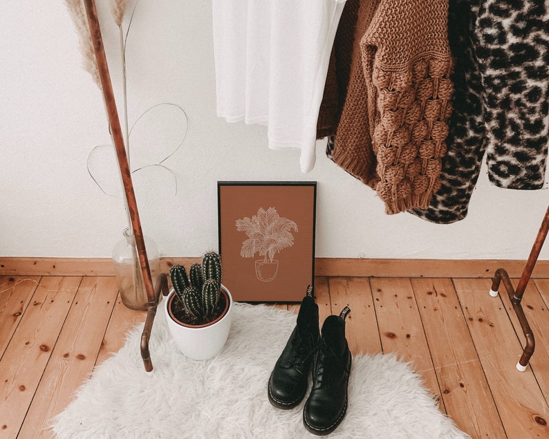 May include: A brown framed print with a line drawing of a palm tree in a pot on a terracotta background. The print is hanging on a white wall above a white fluffy rug with two black leather boots on it.