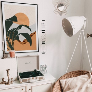 May include: A white record player with a black turntable and a brown and green abstract art print on the wall behind it. The record player is on a white dresser with candles and a white lamp with a white shade on a tripod stand.