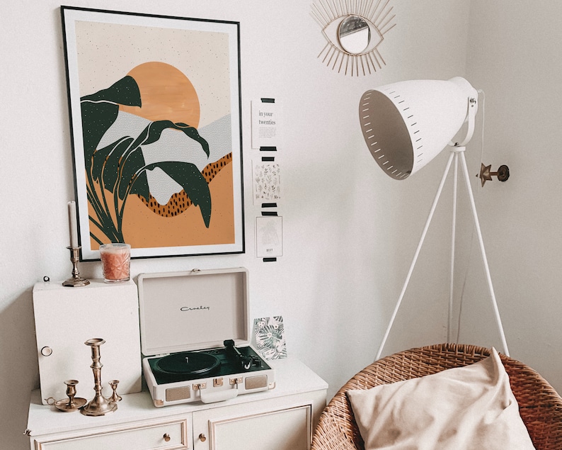 May include: A white record player with a black turntable and a brown and green abstract art print on the wall behind it. The record player is on a white dresser with candles and a white lamp with a white shade on a tripod stand.