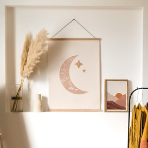 May include: A white wall with two framed prints. The print on the left is a minimalist design of a crescent moon with stars. The print on the right is a landscape scene with mountains and a sun. There are two vases with dried pampas grass on a shelf below the prints.