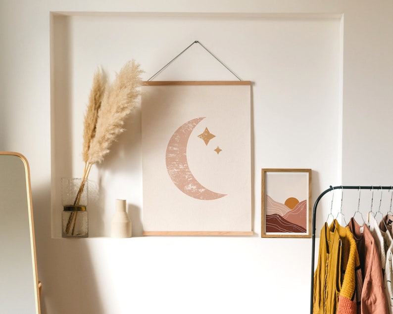 May include: A white wall with two framed prints. The print on the left is a minimalist design of a crescent moon with stars. The print on the right is a landscape scene with mountains and a sun. There are two vases with dried pampas grass on a shelf below the prints.