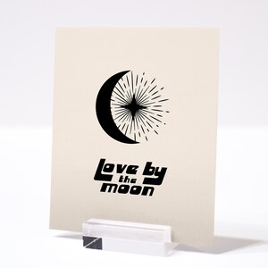 Puede incluir: Una tarjeta beige con un dise&ntilde;o de luna creciente y estrella negros. El texto "Love by the Moon" est&aacute; impreso en negro debajo del dise&ntilde;o.