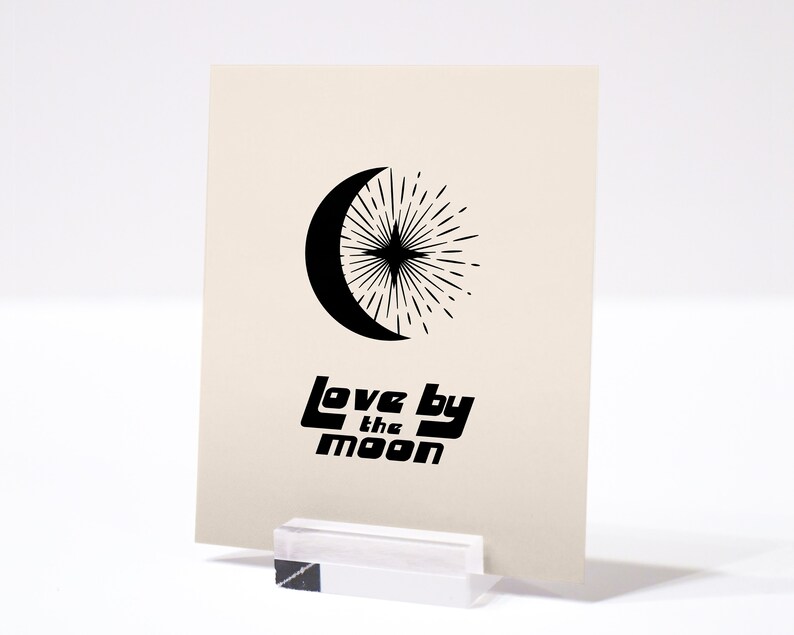 Puede incluir: Una tarjeta beige con un dise&ntilde;o de luna creciente y estrella negros. El texto "Love by the Moon" est&aacute; impreso en negro debajo del dise&ntilde;o.