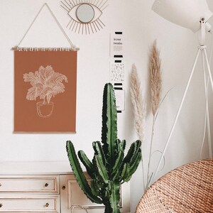 May include: A brown wall with a white framed print of a potted plant, a white lamp with a tripod base, a cactus plant, and a wicker chair. The print has the text "In your twenties" on a black and white striped background.