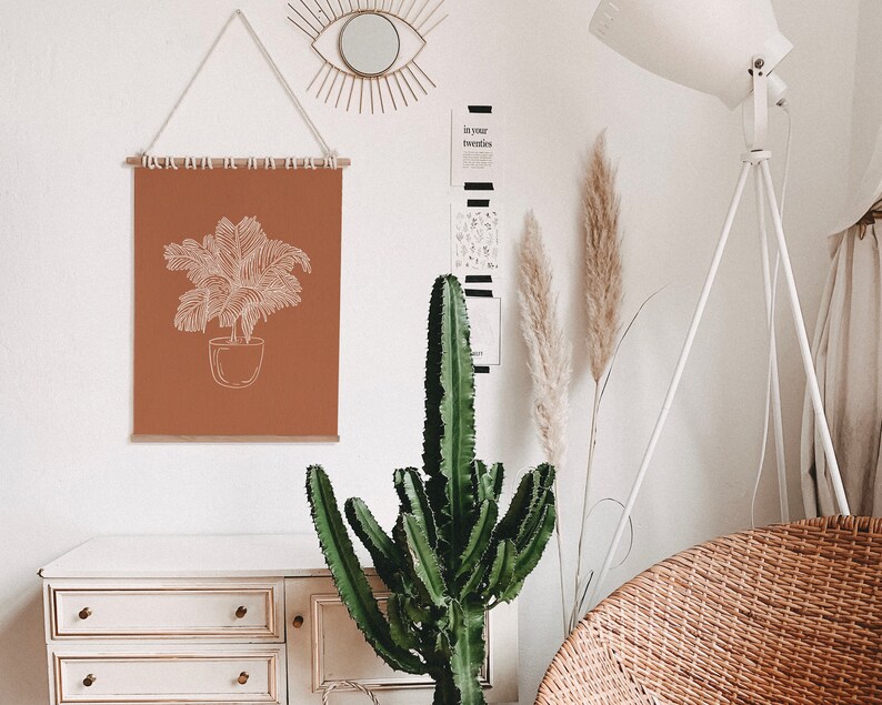 May include: A brown wall with a white framed print of a potted plant, a white lamp with a tripod base, a cactus plant, and a wicker chair. The print has the text "In your twenties" on a black and white striped background.
