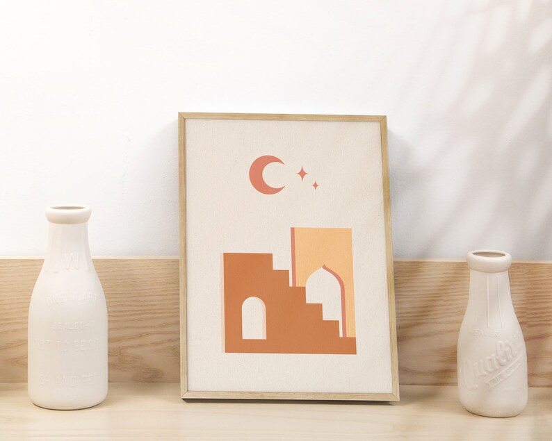 May include: A framed print with a minimalist design featuring a crescent moon and stars above a stylized building with a dome. The building is in shades of orange and brown against a cream background.
