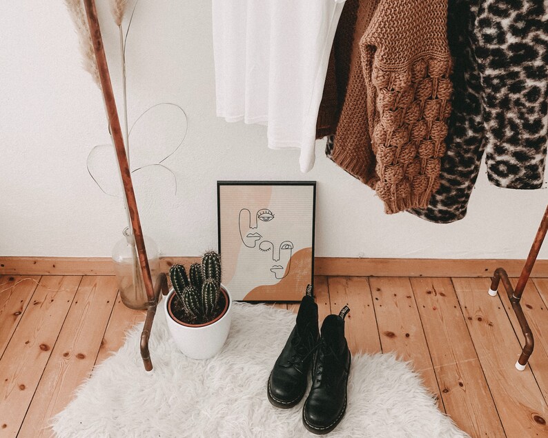 Puede incluir: Una pared blanca con un estante de tuber&iacute;a de cobre que sostiene un su&eacute;ter marr&oacute;n, un su&eacute;ter de estampado de leopardo y una camisa blanca. Una planta de cactus se encuentra sobre una alfombra blanca y esponjosa frente al estante. Una impresi&oacute;n de arte abstracto en blanco y negro cuelga de la pared sobre la alfombra. Dos botas de cuero negras est&aacute;n sobre la alfombra.