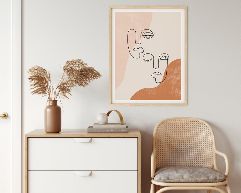 Puede incluir: Una impresi&oacute;n enmarcada con un dibujo minimalista de tres caras en tonos marrones, rosas y blancos. Las caras son abstractas y sin rasgos, solo se dibujan los ojos, la nariz y la boca.