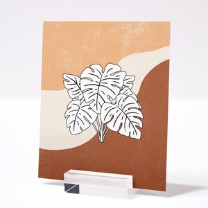 May include: A printable art print featuring a minimalist design of white monstera leaves on a background of brown and orange.