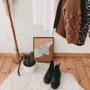 May include: A brown wooden clothes rack with a white shirt, a brown sweater, and a leopard print sweater hanging on it. A potted cactus sits on the floor in front of the rack. A framed print with a brown and white abstract design hangs on the wall behind the rack.