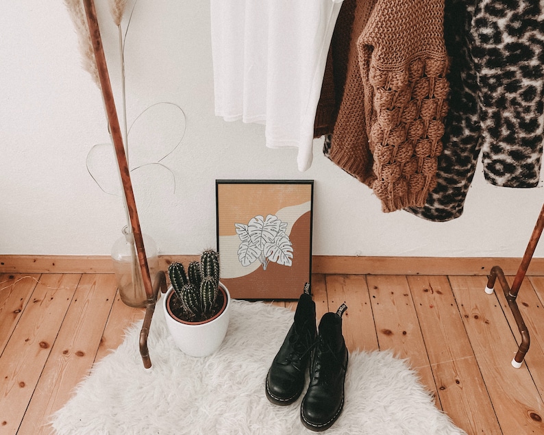 May include: A brown wooden clothes rack with a white shirt, a brown sweater, and a leopard print sweater hanging on it. A potted cactus sits on the floor in front of the rack. A framed print with a brown and white abstract design hangs on the wall behind the rack.